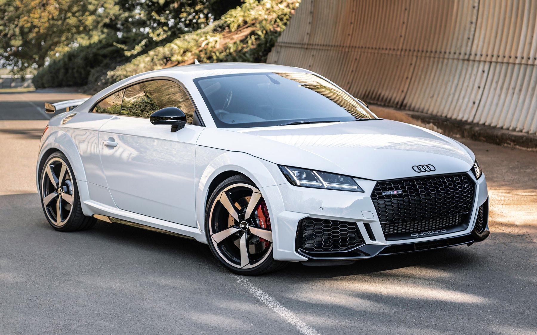 White Audi TT RS 8S coupé with the RS honeycomb grille, quattro script, red brake calipers and fixed rear wing, parked by a corrugated wall