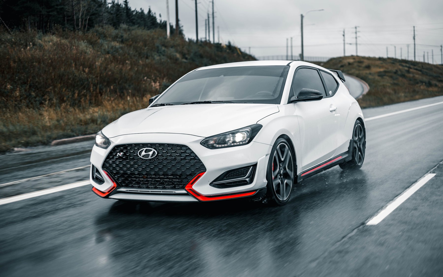 White Hyundai Veloster N with the N grille badge and red lip, driving on a wet road