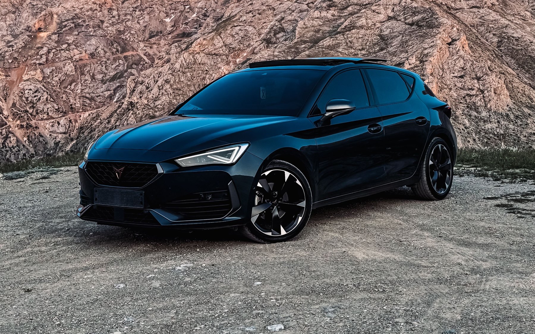 Dark blue Cupra Leon VZ 2.0 TSI hatchback, front three-quarter view, parked on gravel in front of a rocky mountain at dusk