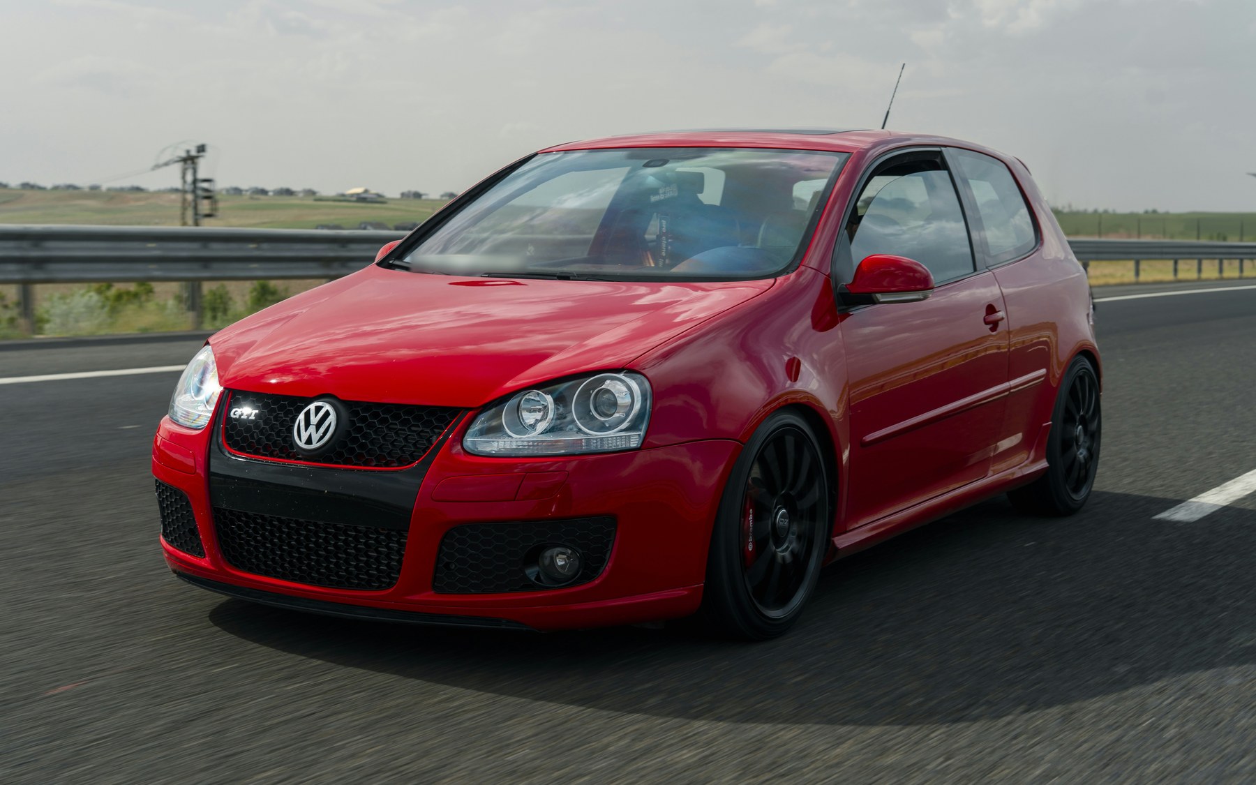 Red three-door VW Golf GTI Mk5 with the GTI badge and red grille outline, driving on a highway